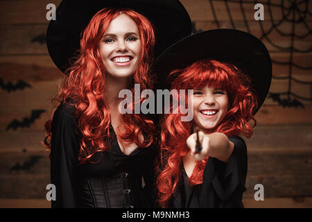 Schönen weißen Mutter und ihre Tochter mit langen roten Haaren in Hexe Kostüme und Zauberstab feiern Halloween posiert mit über Fledermäuse und Spinnennetz auf Holz- studio Hintergrund. Stockfoto