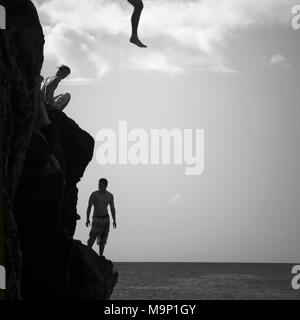 Silhouette junge Männer und das Bein eines Mannes, der mitten in der Luft, weg von der 'Da Big Rock' oder 'Jump Rock', ein Felsen, der sich perfekt in der Bucht von Waimea Bay Beach Park positioniert ist, O'ahu, Hawaii. Stockfoto