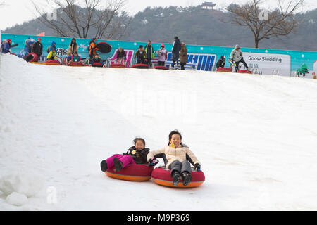Eine asiatische Frau und ihre Tochter Spaß Schlittenfahren auf einem steilen Eis Hang. Die Hwacheon Sancheoneo Ice Festival ist eine Tradition für die Menschen in Korea. Jedes Jahr im Januar Menschenmassen versammeln sich auf dem zugefrorenen Fluss der Kälte und dem Schnee des Winters zu feiern. Hauptattraktion ist Eisfischen. Jung und Alt warten geduldig auf ein kleines Loch im Eis für eine Forelle zu beißen. In zelten Sie können den Fisch vom Grill, nach dem sie gegessen werden. Unter anderem sind Rodeln und Eislaufen. Die in der Nähe Pyeongchang Region wird Gastgeber der Olympischen Winterspiele im Februar 2018. Stockfoto