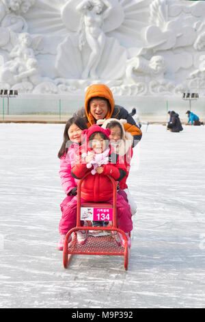 Eine asiatische Familie, die auf eine besondere Gruppe Schlitten an der Hwacheon Sancheoneo Ice Festival. Die Hwacheon Sancheoneo Ice Festival ist eine Tradition für die Menschen in Korea. Jedes Jahr im Januar Menschenmassen versammeln sich auf dem zugefrorenen Fluss der Kälte und dem Schnee des Winters zu feiern. Hauptattraktion ist Eisfischen. Jung und Alt warten geduldig auf ein kleines Loch im Eis für eine Forelle zu beißen. In zelten Sie können den Fisch vom Grill, nach dem sie gegessen werden. Unter anderem sind Rodeln und Eislaufen. Die in der Nähe Pyeongchang Region wird Gastgeber der Olympischen Winterspiele im Februar 2018. Stockfoto