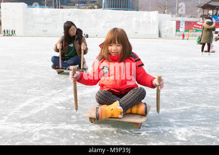 Zwei Mädchen sich nach vorn drücken, auf Schlitten an der Hwacheon Sancheoneo Ice Festival. Die Hwacheon Sancheoneo Ice Festival ist eine Tradition für die Menschen in Korea. Jedes Jahr im Januar Menschenmassen versammeln sich auf dem zugefrorenen Fluss der Kälte und dem Schnee des Winters zu feiern. Hauptattraktion ist Eisfischen. Jung und Alt warten geduldig auf ein kleines Loch im Eis für eine Forelle zu beißen. In zelten Sie können den Fisch vom Grill, nach dem sie gegessen werden. Unter anderem sind Rodeln und Eislaufen. Die in der Nähe Pyeongchang Region wird Gastgeber der Olympischen Winterspiele im Februar 2018. Stockfoto