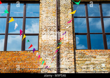 Bunte dreieckige Dekorationsfahnen hängen auf dem Hintergrund der alten loft Backstein Gebäude mit großen Fenstern. Hintergrund Stockfoto