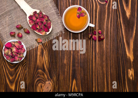 Flach Layout mit Kräutertee mit rosenblütenblätter. Weiße Tasse mit heißem Kaffee. Ein Löffel aus Holz mit getrockneten Blütenknospen auf einer Leinwand Serviette. Ansicht von oben Stockfoto