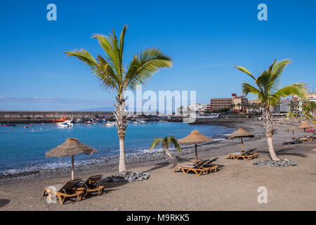 Palmen, Sonnenliegen und Stroh Sonnenschirm Sonnenschirme am Strand mit Blick auf die Boote im Hafen in Playa San Juan, Teneriffa, Kanarische Inseln, Spanien Stockfoto