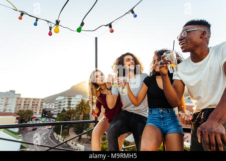 Vier junge Leute trinken und lächelnd auf dem Dach in Abend. Männer und Frauen feiern mit Getränke auf der Terrasse. Stockfoto