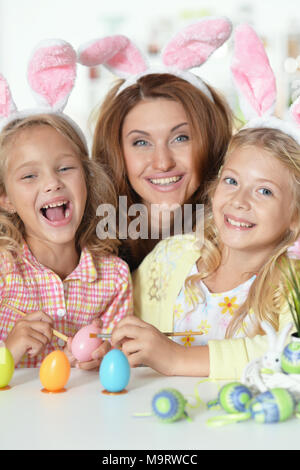 Glückliche junge Mutter mit ihren süßen Töchtern tragen Hasenohren und Bemalen von Ostereiern zu Hause Stockfoto