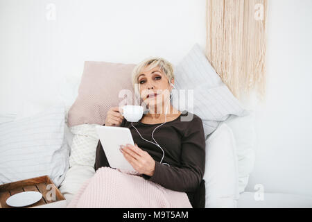 Eine positive Frau trinkt einen Kaffee am Morgen im Bett lag, und verwendet ein Tablet Videos ansehen, Musik hören und mit Freunden über soziale Netzwerke chat Stockfoto