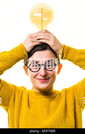 Inspiration und Kreativität - die Glühbirne Moment: Ein junger "geeky" Kaukasischen Frau Mädchen mit einem gelben Pullover Pullover und der runden Brille, mit einem alten Retro-Stil elektrischen Glühbirne, vor einem weißen Hintergrund, Großbritannien Stockfoto