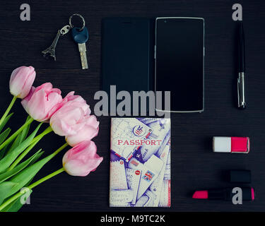 Frau Rosa und Schwarz Zubehör auf dunklem Hintergrund und Handy mit leerer Bildschirm angezeigt. Blumenstrauß aus rosa Tulpen. Travel Concept. Flach. Stockfoto