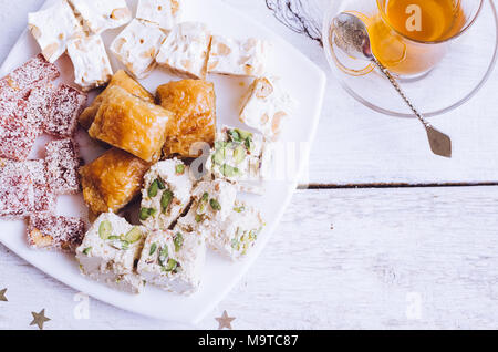 Verschiedene traditionelle östliche Desserts und Tasse Tee auf einem weißen Holz- Hintergrund. Baklava, Halva, rahat Lokum, Sorbet, Nüsse, Datteln, kadayif auf Platte Stockfoto