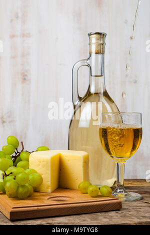 Weißer Wein, Käse und Trauben auf einem alten Holztisch. Wein wird in Glas gegossen Stockfoto