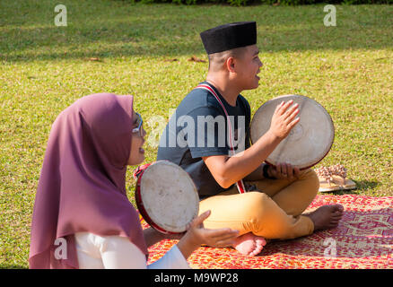Zwei Malaysier, ein Mann mit einem sonkok und eine Frau trägt ein Kopftuch, sitzen auf einer traditionellen rattan Matte und spielen Die kompang (Malaiisch Trommel). Stockfoto