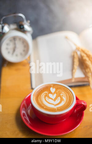 Eine Tasse Latte Kaffee mit offenem Buch und Wecker mit Blume auf dem Tisch im Café Stockfoto