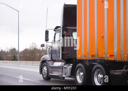 Tag cab Dunkelbraun Big Rig halb LKW-Flotte mit abgedeckten Rahmen orange Sattelschlepper auf der Autobahn bei Regen Wetter für die rechtzeitige Lieferung der Industria Stockfoto