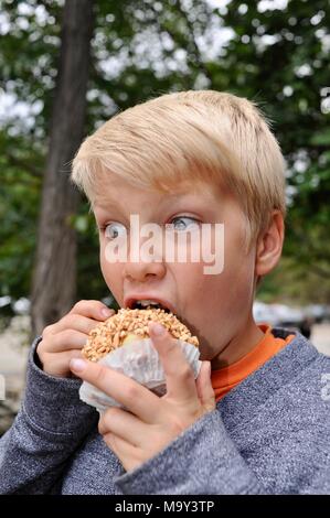 Blonde Junge mit großen Bissen in Erdnuss abgedeckt caramel Apple außerhalb im Herbst am historischen 1837 Franklin Cider Mill, Bloomfield Hills, Michigan, USA. Stockfoto