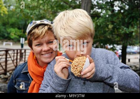 Blonde Junge mit großen Bissen in Erdnuss abgedeckt caramel Apple außerhalb im Herbst am historischen 1837 Franklin Cider Mill, Bloomfield Hills, Michigan, USA. Stockfoto