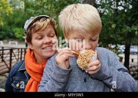 Blonde Junge mit großen Bissen in Erdnuss abgedeckt caramel Apple außerhalb im Herbst am historischen 1837 Franklin Cider Mill, Bloomfield Hills, Michigan, USA. Stockfoto