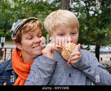 Blonde Junge mit großen Bissen in Erdnuss abgedeckt caramel Apple außerhalb im Herbst am historischen 1837 Franklin Cider Mill, Bloomfield Hills, Michigan, USA. Stockfoto