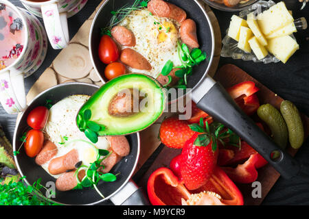 Deutsch Gesundheit Frühstück für zwei. Spiegeleier mit Würste in einer Pfanne mit Gewürzen und frischem Gemüse, Käse, Nüsse, schwarz und Obst Tasse Tee. Stockfoto