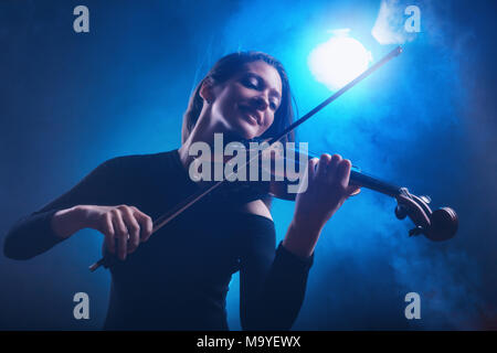 Schöne junge Frau spielt die Geige auf dunkelblauem Hintergrund. Nebel im Hintergrund. Studio shot Stockfoto