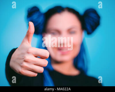 Enthusiastisch motivierten attraktive junge Frau mit blauen Zöpfe Frisur geben einen Daumen nach oben Geste der Zustimmung und der Erfolg mit einem strahlenden Lächeln. Fokus auf der Hand. Studio gedreht. Stockfoto