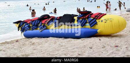 Gelbe aufblasbare Banane mit Paddeln auf der belebten Strand mit Menschen und Boote für den Hintergrund. Stockfoto