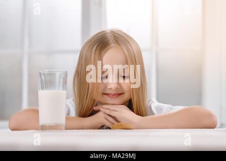 Ein wunderschönes kleines Mädchen sitzt in der Nähe von Tisch, mit weißen Tischtüchern bedeckt. Es ist transparent Glas mit Milch. Kind trägt weiße Sweatshirt und hat nettes Lächeln. Sie hat gerade lange blonde Haare. Stockfoto