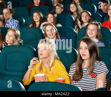 Verlassen girls watching Film im Kino. Kinder ooking sehr interessiert, Trinken kohlensäurehaltige Getränke, sitzen auf den Hintergrund der anderen Kinder. Sie tragen schicke bunte Kleidung. Suchen Angst. Stockfoto