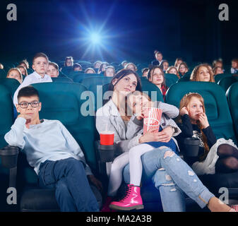 Ziemlich ältere Schwester schaut Film oder ein Cartoon in der neuen Kinosaal mit Junior Kinder. Kinder sehr interessiert und verlassen. Es gibt auch andere Menschen auf der backgroung. Stockfoto