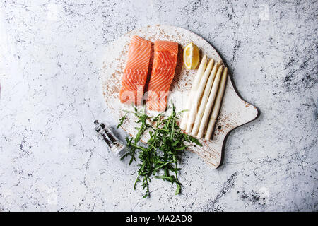 Geschnittenem rohen ungekochten Lachs filet auf Holz Schneidebrett als Fisch mit Zutaten zum Kochen Abendessen Rucola, weißer Spargel über Weiß marbl Stockfoto