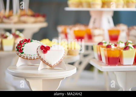 Runde und herzförmige glasierte Plätzchen, mit Glasur Blumen und Muster verziert sind auf dem Stand aus Holz im Restaurant. Es gibt eine bunte leckere Schokoriegel hinter Ihnen. Gute Wahl für eine Hochzeit. Stockfoto