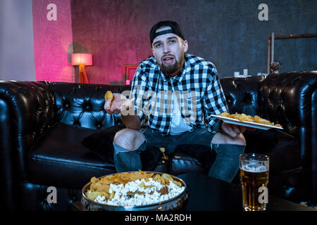 Junge fanatischer Mann, Fußball Spiel auf Fernsehen nervös und aufgeregt leiden Stress auf Sofa zu Hause mit Ball Bier Flasche und Chips aussehen Stockfoto