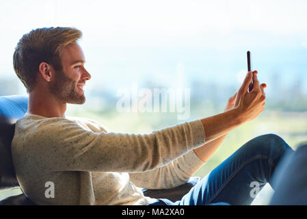 Profil Bild von einem gutaussehenden jungen kaukasischen Mann lächelnd, während Sie einen selfie mit seinem Handy, mit einem wunderschönen Blick auf die Stadt in der Dist Stockfoto