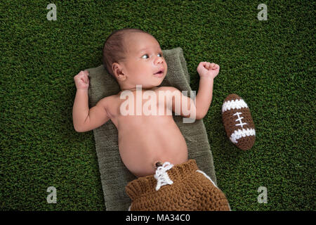 2 Woche alt, Alert, neugeborenes Baby Boy liegen auf dem Rasen Rasen mit einem gehäkelten American Football. Stockfoto