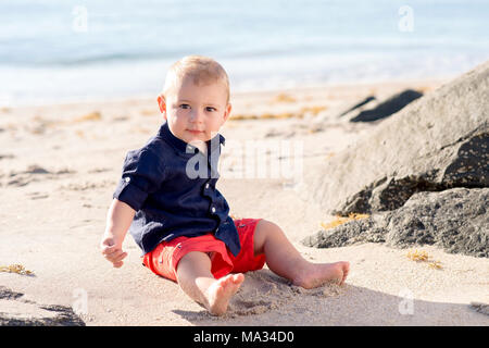 Ein 1 Jahr altes Baby Junge sitzt auf einem Strand und direkt auf die Kamera. Stockfoto