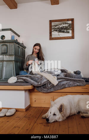 Junge Frau sitzt neben einem Kachelofen und strickt, Hund auf dem Boden liegt Stockfoto