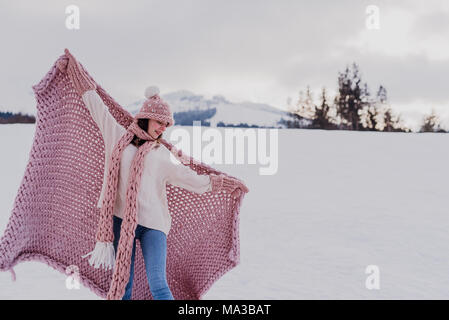 Junge Frau steht mit rosa Decke, wolle Mütze und Schal im Schnee, breitete die Arme, Stockfoto