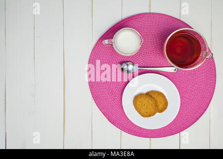 Heißen Tee, Kekse und Milch sind auf einem rosa Tischset auf eine weiße Tafel Tabelle angeordnet. Die Anordnung ist außermittig und Schuß von Overhead. Stockfoto