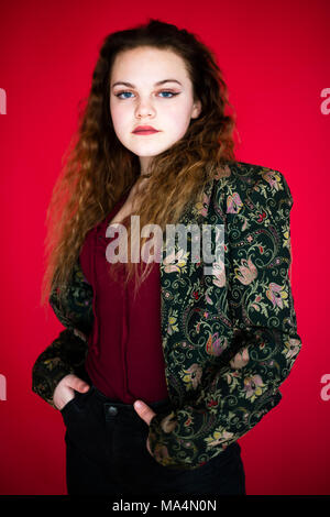 Moody's zu finden. Einen Motivationsanreiz Jungen 13 13 Jahre alten Teenager jugendliche Mädchen mit langen roten Haaren auf rotem Hintergrund, in einem Fotostudio, Großbritannien Stockfoto