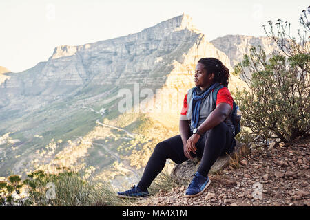 Afrikanische Wanderer eine Pause neben dem Wanderweg auf einem bergigen Trail zu nehmen in die fantastische Aussicht auf die Berge um ihn herum, die Stockfoto