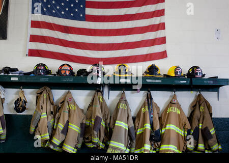 Schutzkleidung, Helme der Feuerwehrleute, und Wahlbeteiligung Mäntel zusammen mit einer amerikanischen Flagge auf Anzeige in der örtlichen freiwilligen Firehouse Stockfoto