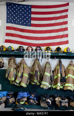 Schutzkleidung, Helme der Feuerwehrleute, und Wahlbeteiligung Mäntel zusammen mit einer amerikanischen Flagge auf Anzeige in der örtlichen freiwilligen Firehouse Stockfoto