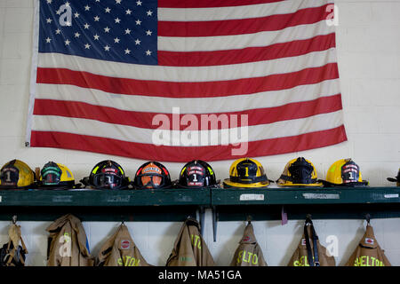 Schutzkleidung, Helme der Feuerwehrleute, und Wahlbeteiligung Mäntel zusammen mit einer amerikanischen Flagge auf Anzeige in der örtlichen freiwilligen Firehouse Stockfoto