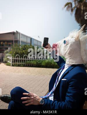 Junge ungewöhnliche Mann in Pferd Kopf Maske und eleganten Anzug macht ein Foto per Telefon. Einhorn ist Genießen Sie warmen Sommertag. Merkwürdige Kerl sitzt auf der Werkbank Stockfoto