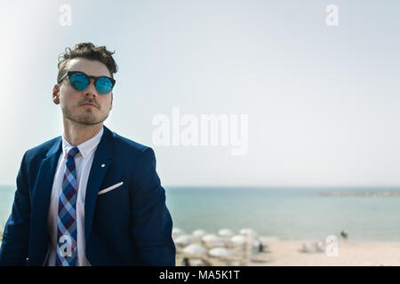 Junge ernster Mann im eleganten Anzug und Sonnenbrille auf dem Hintergrund von Meer und Himmel. Erfolgreiche Manager ist Entspannung im Freien in sonniger Tag. Trendy Kerl Stockfoto