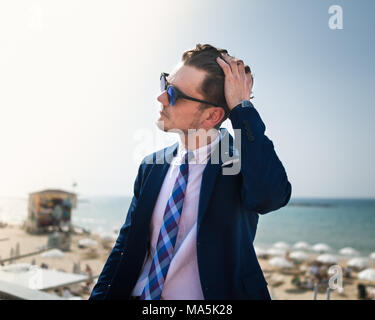 Junger Mann korrigiert Haar auf dem Hintergrund von Meer und Himmel. Erfolgreicher Geschäftsmann in Anzug und Brille ist im Freien in sonniger Tag. Trendy Kerl Stockfoto