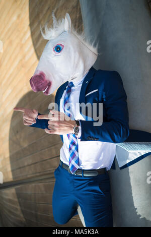 Junge cool Mann in Anzug und lustige Maske aktiv gestikulieren. Ungewöhnliche Manager verweist auf Sie. Einhorn breitet Hände an der Promenade und hat auf der Bank entspannen Stockfoto