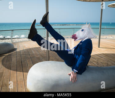 Junger Mann in Anzug fällt unter Regenschirmen auf Stadt am Wasser. Ungewöhnliche Geschäftsmann in lustige Maske City Promenade. Unicorn in komischen Pose Stockfoto