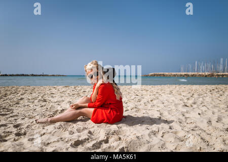 Junge Frau im roten Kleid sitzt am Strand. Blonde Mädchen sitzt auf dem Hintergrund von Meer und Himmel. Weibliche Reisende entspannt am Strand