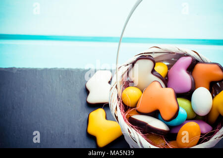 Kleine weiße Korb voller Süßigkeiten Ostereier und Hasen Pastellfarben auf Schiefer und blau Holz- Hintergrund. Copyspace Stockfoto